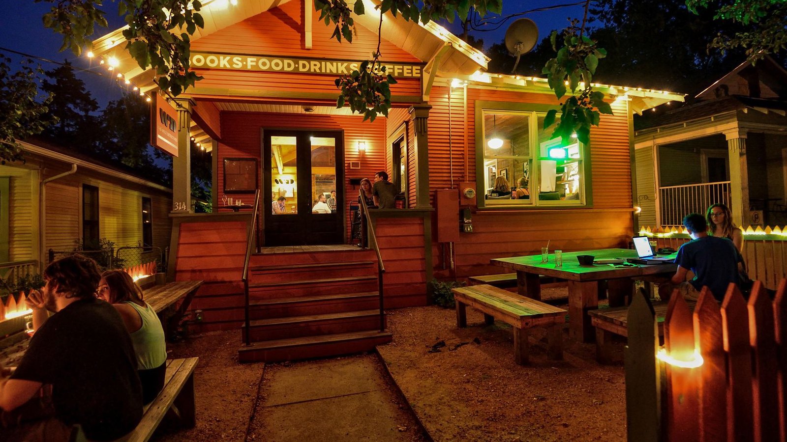 People sitting together at outdoor tables outside a warmly lit bookshop in the evening